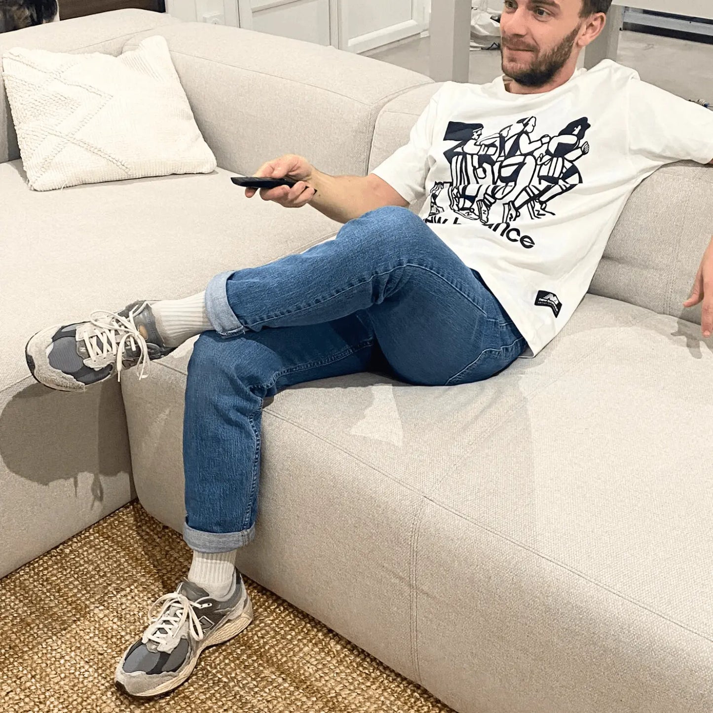 Un homme sur son canapé avec une télécommande à la main, portant un t-shirt blanc New Balance, un jean bleu et une paire de New Balance 2002R Protection Pack Rain Cloud