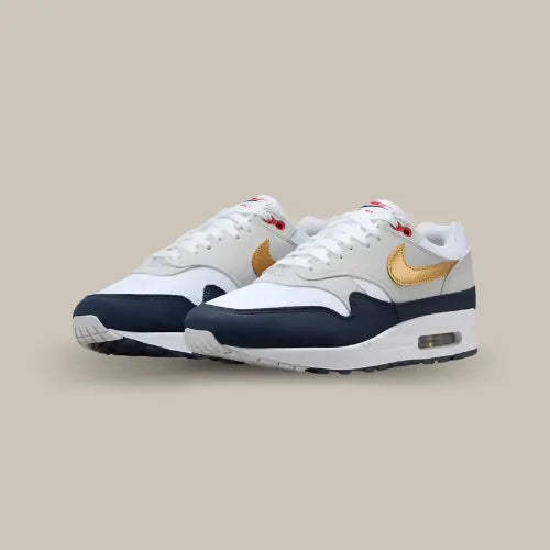 Nike Air Max 1 Olympic vue en angle trois quarts, paire de sneakers blanche avec empiècements bleu marine, Swoosh doré et détails rouges, design rétro-running avec unité Air iconique.