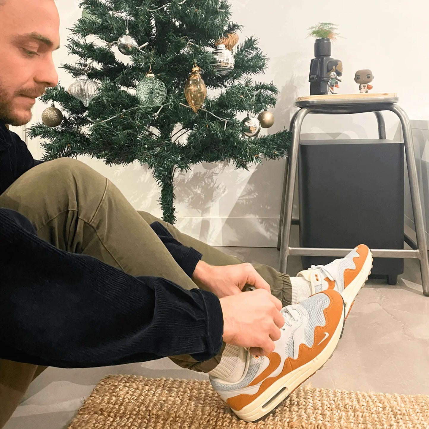 Un homme devant un sapin de noel habillé d'une veste noir et d'un pantalon vert kaki laçant ses Nike Air Max 1 Patta « Monarch »