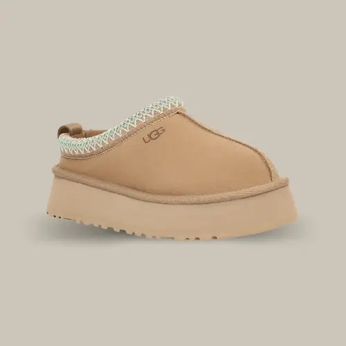La UGG Tazz Slipper Sand vue de côté avec sa couleur sable, son épaisse plateforme de 3.8 cm et sa tresse brodée blanche.