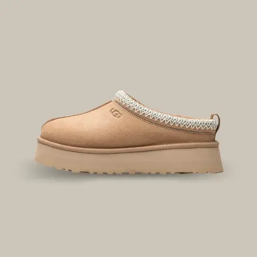 La UGG Tazz Slipper Sand vue de côté avec sa couleur sable, son épaisse plateforme de 3.8 cm et sa tresse brodée blanche.
