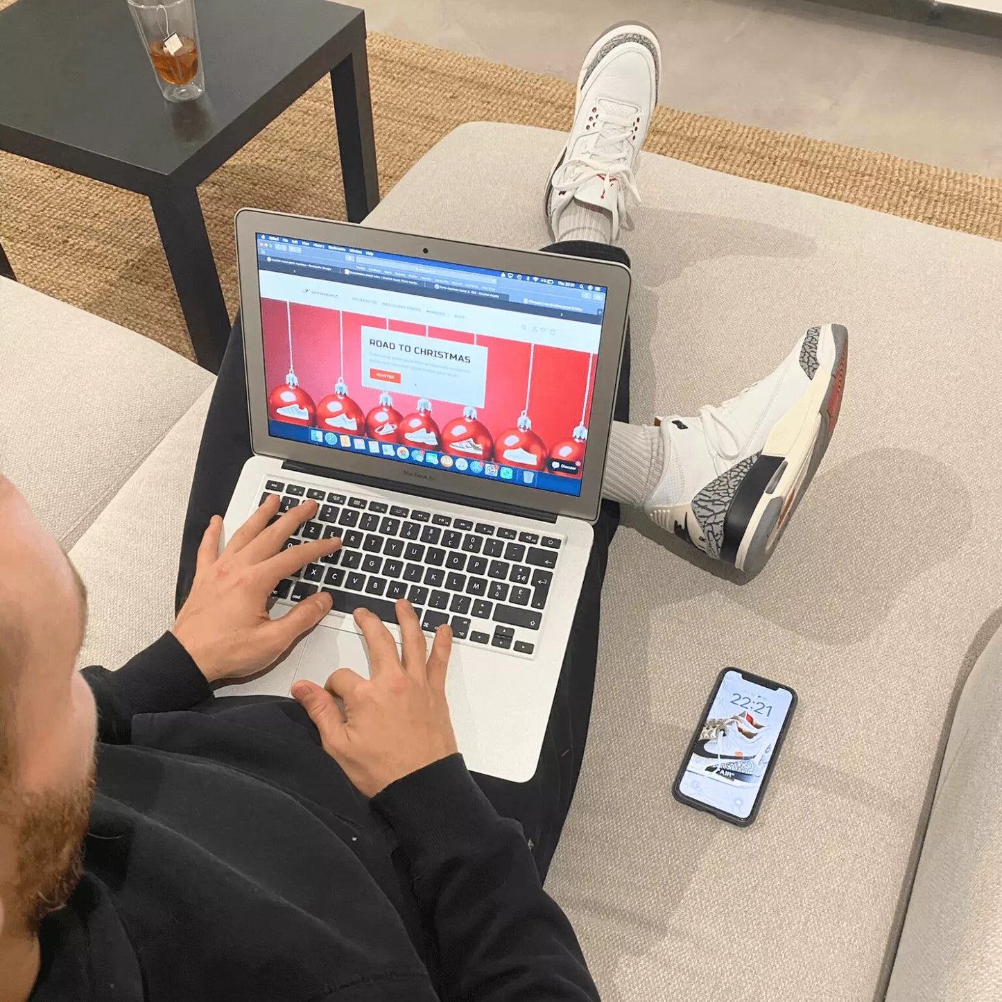 Un homme sur un canapé avec un macbook dans les mains ouvert sur la page d'accueil du site OFFSNEAKZ et portant aux pieds une paire de Jordan 3 retro white ciment reimagined