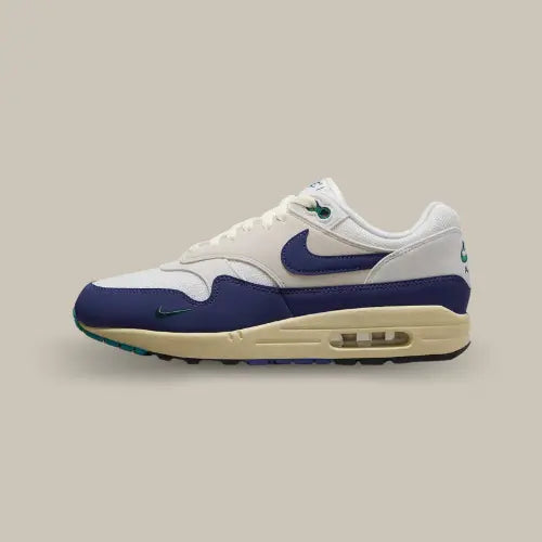 Nike Air Max 1 Athletic Department Deep Royal Blue vue latérale, sneaker en cuir et mesh blanc cassé avec empiècements bleu royal et mini Swoosh vert, design rétro running et unité Air visible.
