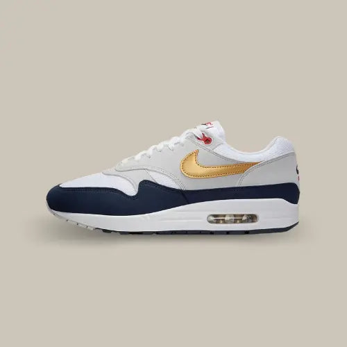 Nike Air Max 1 Olympic vue latérale, sneaker blanche avec empiècements gris et bleu marine, Swoosh doré métallique et unité Air visible, design inspiré des Jeux Olympiques.
