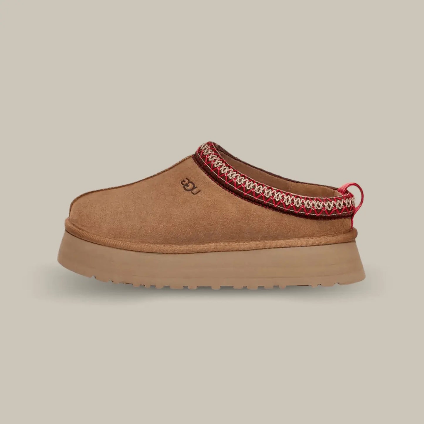 La UGG Tazz Slipper Chestnut avec une tige en daim, une tresse rouge et blanche en polyester recyclé et sa semelle compensée en EVA de 3.8cm.