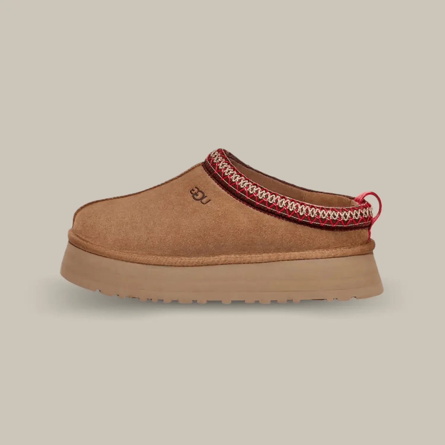 La UGG Tazz Slipper Chestnut avec une tige en daim, une tresse rouge et blanche en polyester recyclé et sa semelle compensée en EVA de 3.8cm.