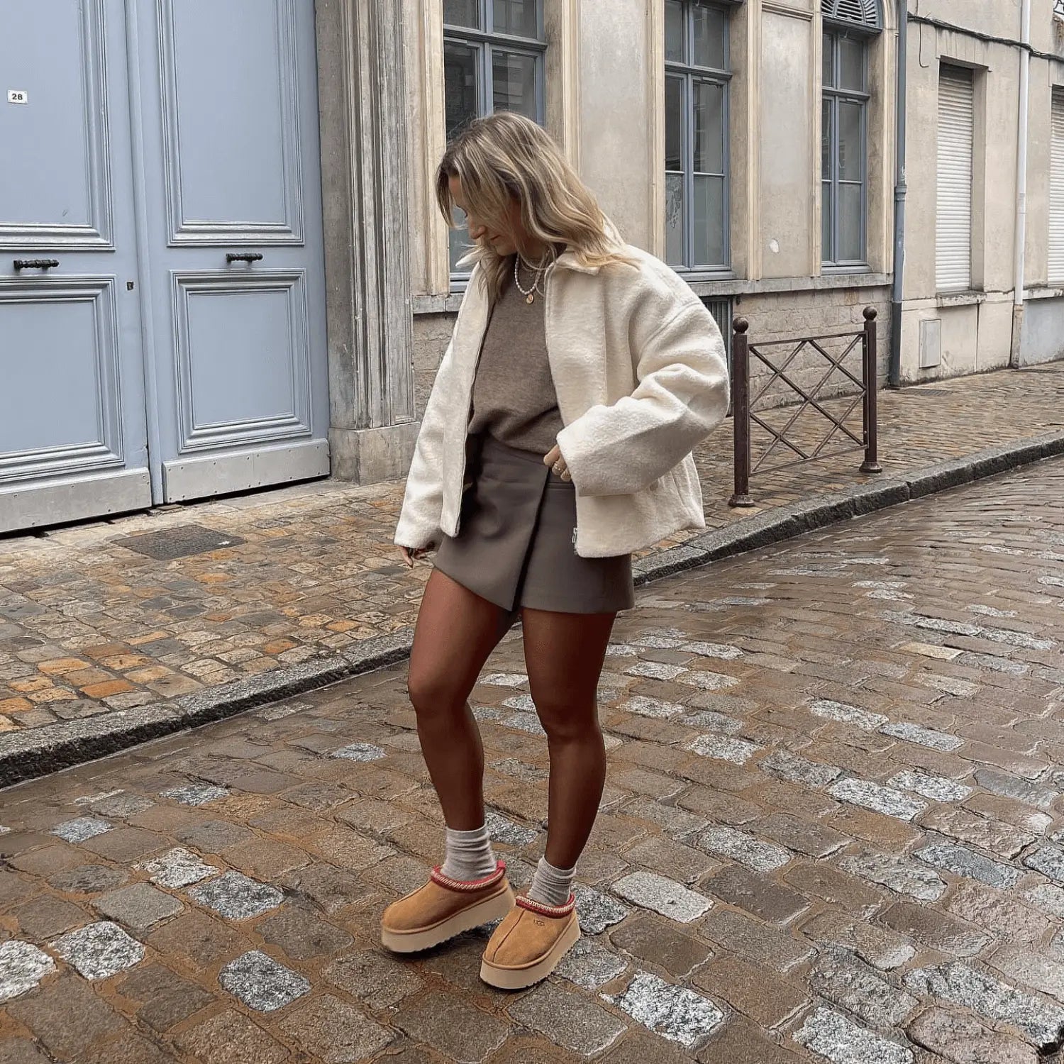 Modèle femme photographié dans la rue portant une paire de UGG Tazz Chestnut avec une jupe grise un pull marron et une veste beige 